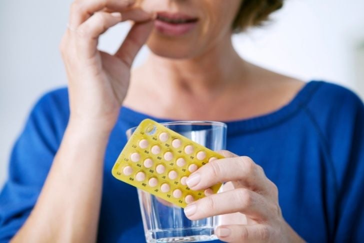 Image shows woman holding a glass and pill packet in one hand while she puts a pill in her mouth with the other hand
