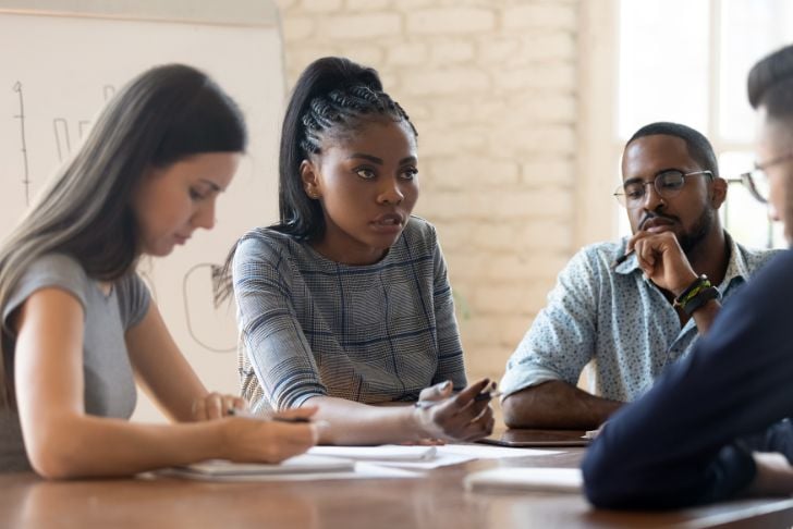Image shows a diverse group of people at a table in discussion while one woman takes notes