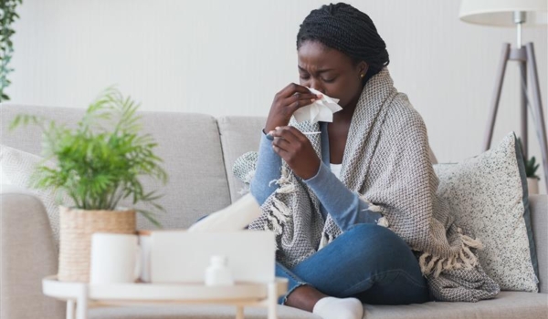 Image shows young woman sitting cross-legged in a sofa with a shawl around her shoulders holding a tissue to her nose