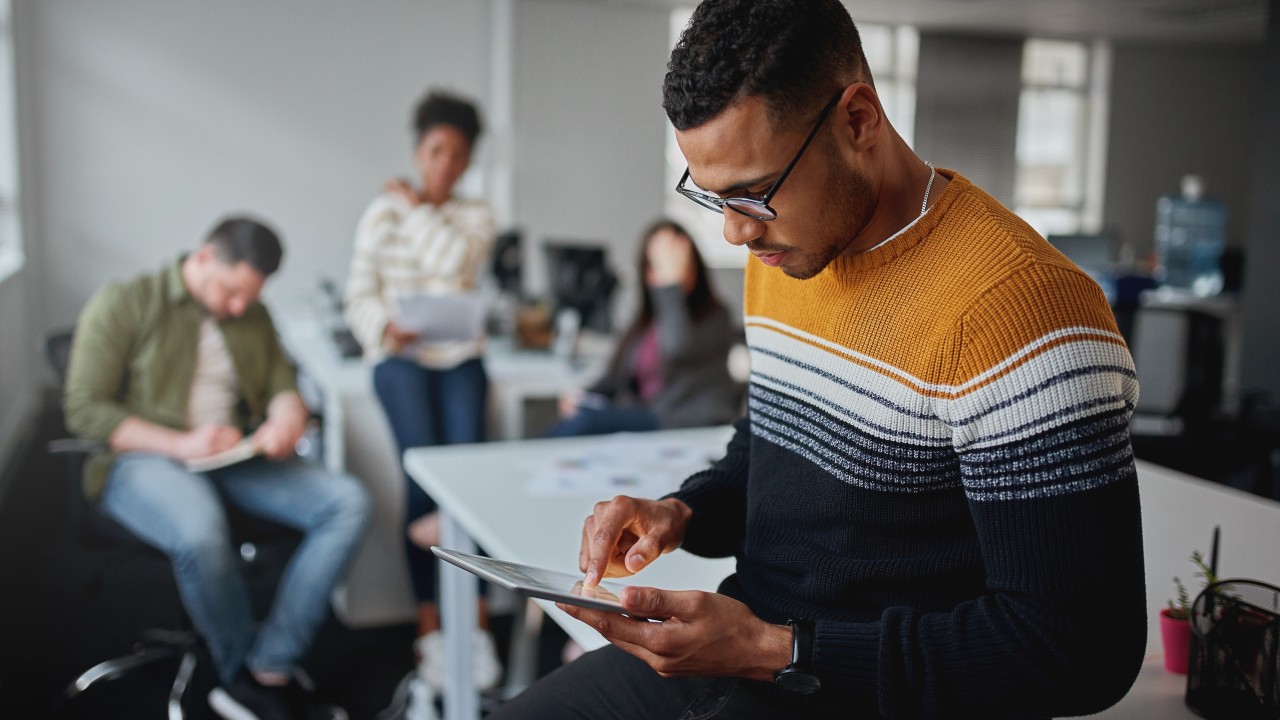 Employee using a tablet in a workplace setting while colleagues collaborate in the background, representing different working styles and workplace inclusion.