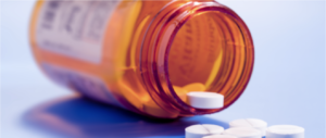 Image shows an open pill bottle on its side with one pill balanced on the lip and a few on the surface beside the pill bottle