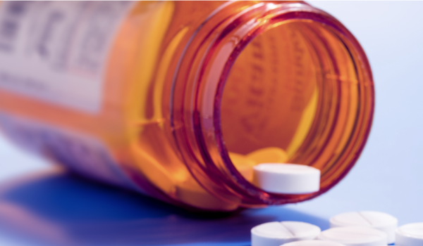 Image shows an open pill bottle on its side with one pill balanced on the lip and a few on the surface beside the pill bottle