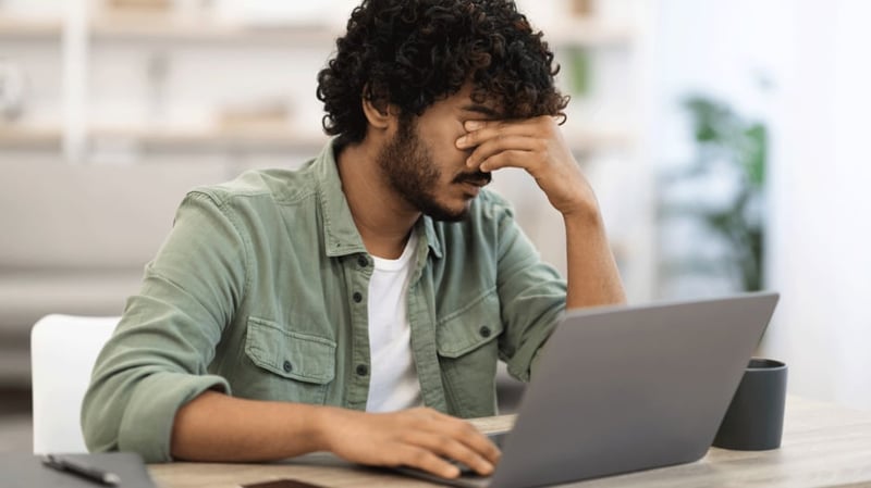 Man rubs his eyes while sat at a desk with a laptop on it