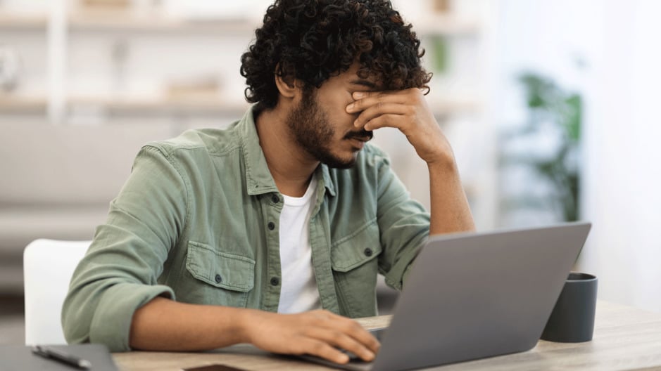 Man rubs his eyes while sat at a desk with a laptop on it