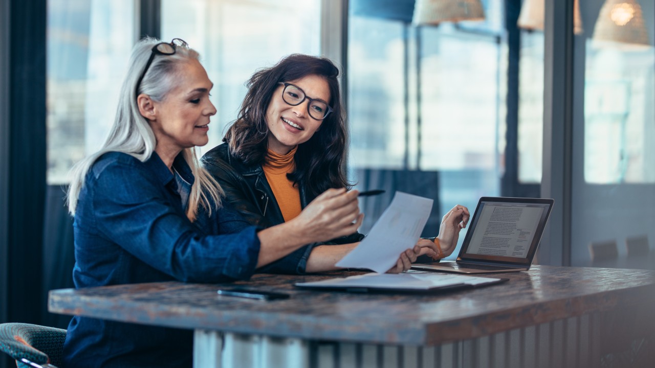 Two colleagues discussing documents at a table with a laptop.
