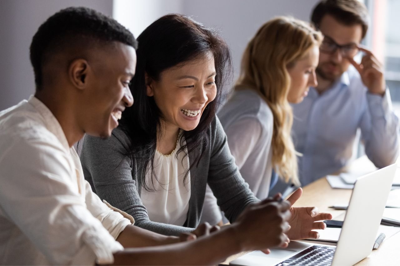 Image shows people working in an office side by side with two colleagues smiling at a laptop