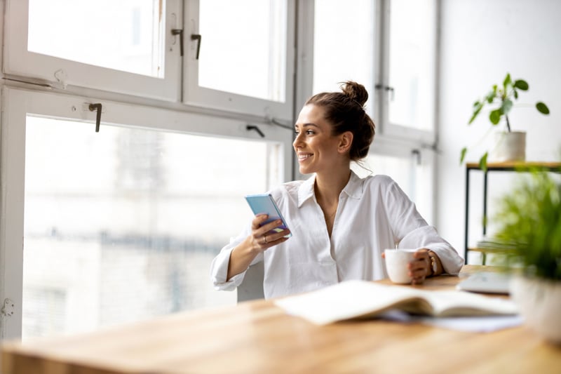 Woman sitting next to a desk looking out the window holding her phone