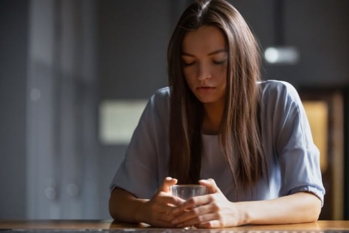 Image shows a young woman sitting at a table with a glass in her hands