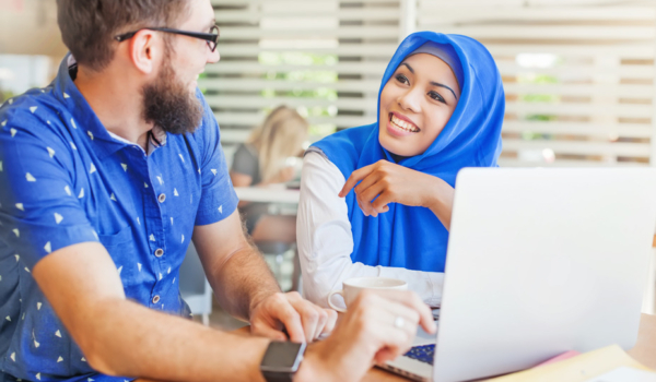 Image shows a young man and woman smiling at each other behind a laptop