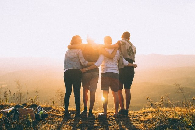 Image shows four people from behind with their arms around each other's shoulders as they look down from a hill's edge