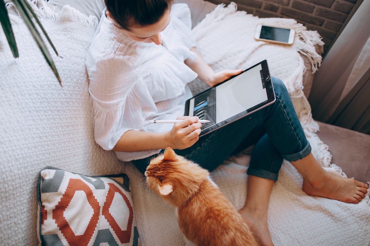 Woman on a sofa using a tablet with her ginger cat beside her